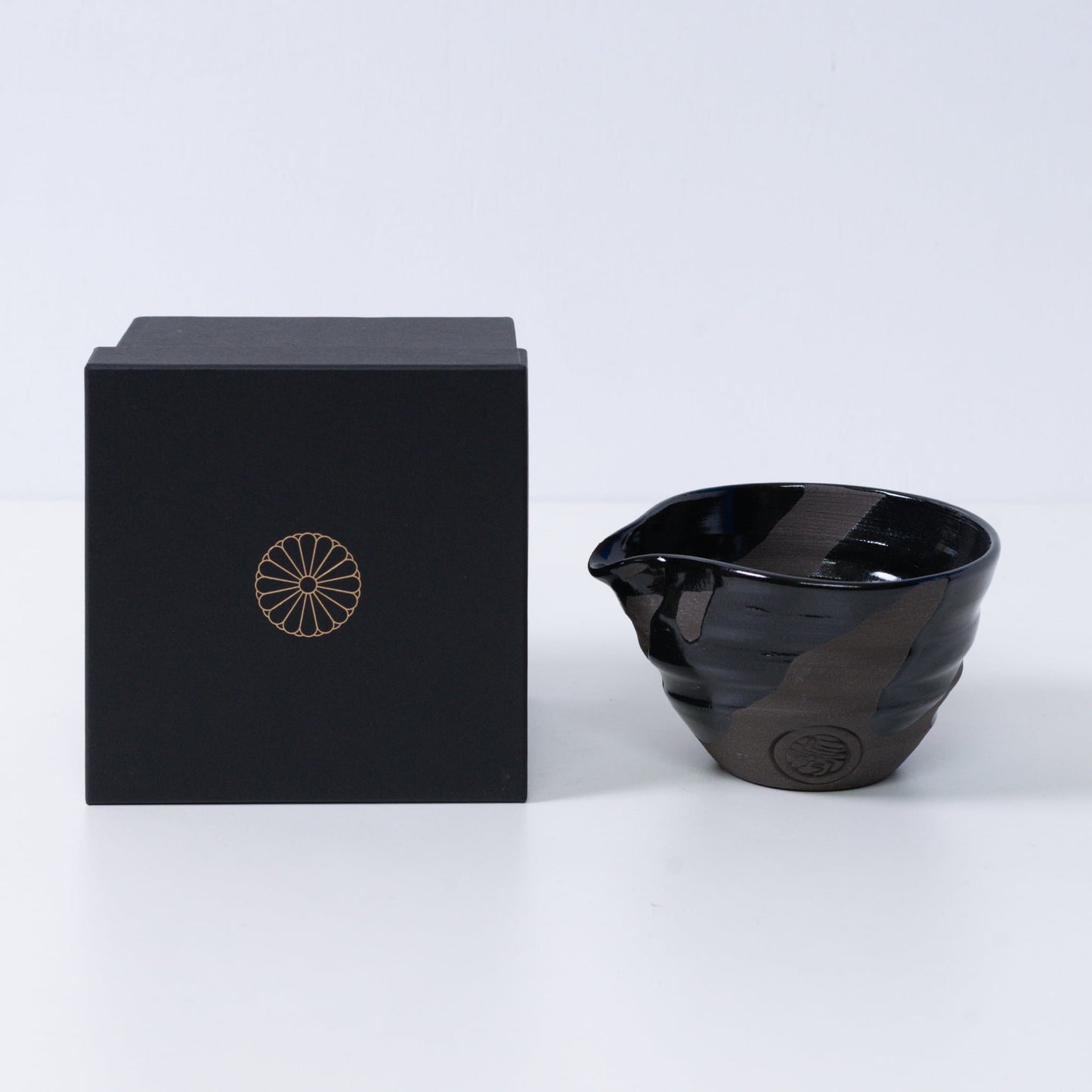 Japanse matchakom met tuit "Sumi Black" aardewerk naast een zwarte geschenkdoos met een gouden keizerlijke chrysant-embleem, keramiek uit Kyoto.