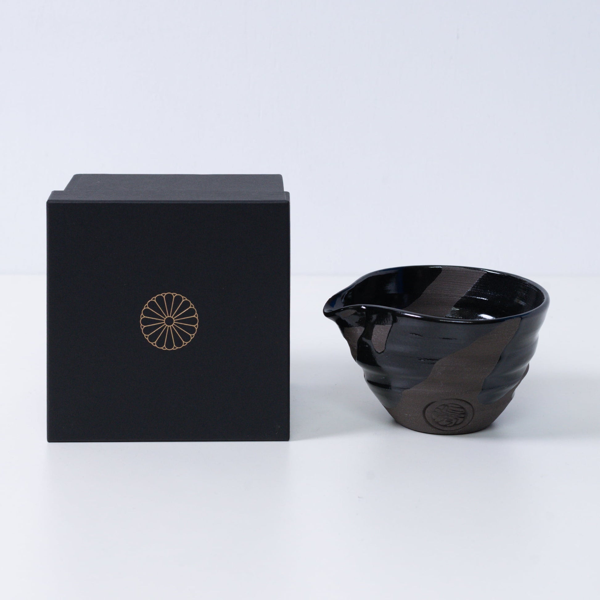 Japanse matchakom met tuit "Sumi Black" aardewerk naast een zwarte geschenkdoos met een gouden keizerlijke chrysant-embleem, keramiek uit Kyoto.
