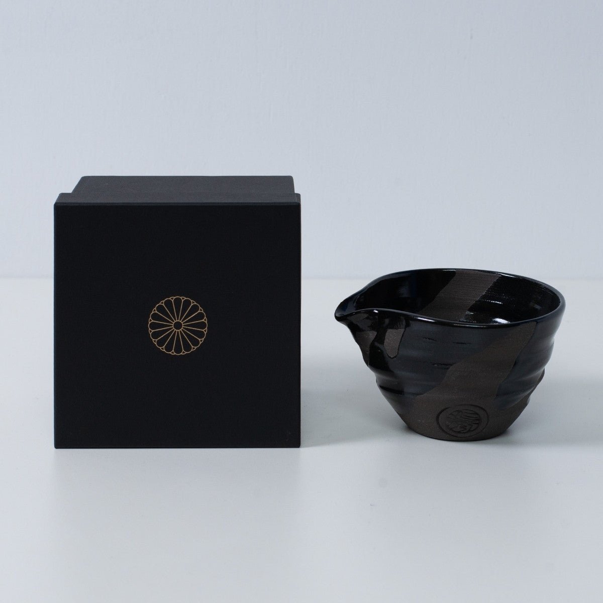 Japanese matcha bowl with spout "Sumi Black" pottery next to a black gift box with a gold imperial chrysanthemum crest, Kyoto ceramics.
