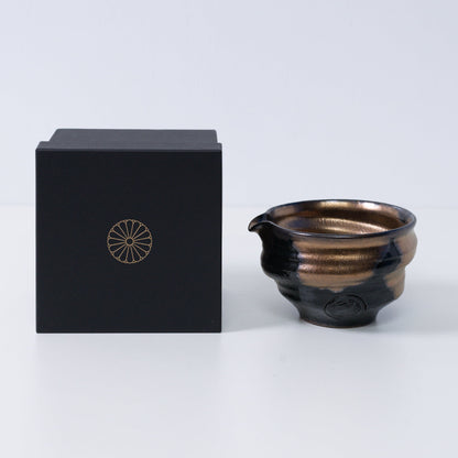 Gouden en zwarte Japanse matchakom met tuit "Zen" aardewerk naast een zwarte geschenkdoos met een gouden chrysant-embleem, gemaakt in Japan.