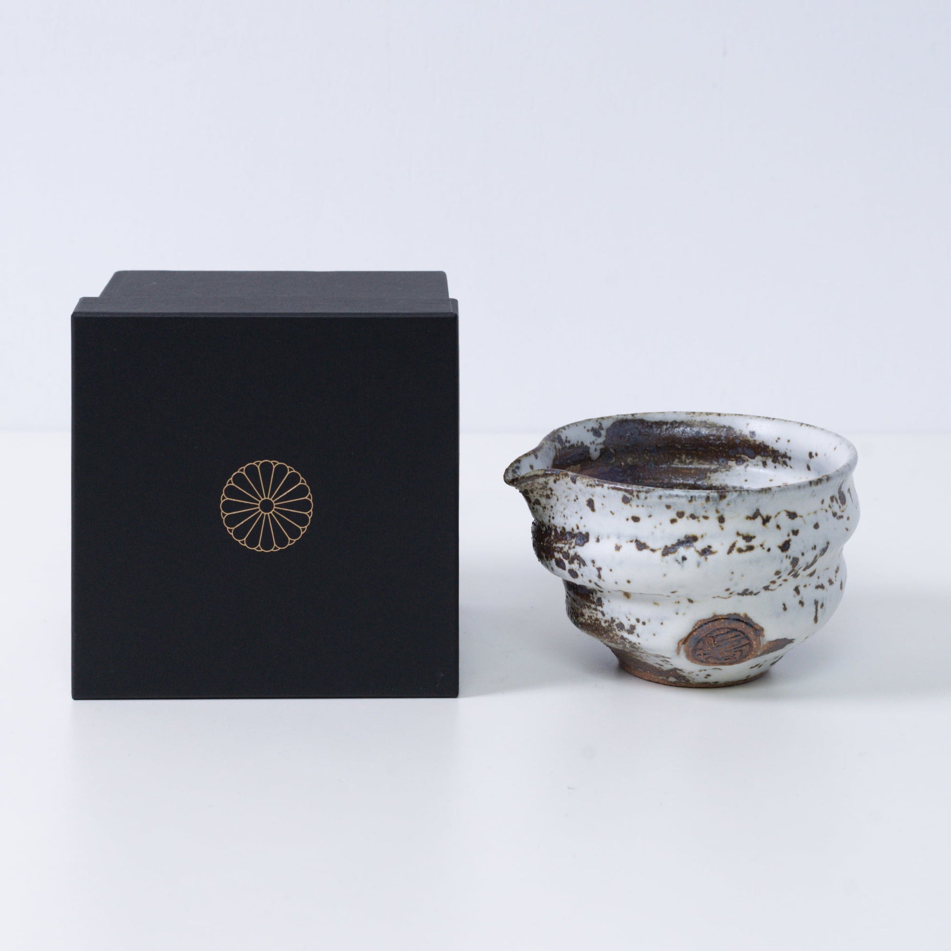 Bruine en witte Japanse matchakom met tuit, "Zui" aardewerk, naast een zwarte geschenkdoos met een gouden keizerlijke chrysant-embleem, uit Japan.