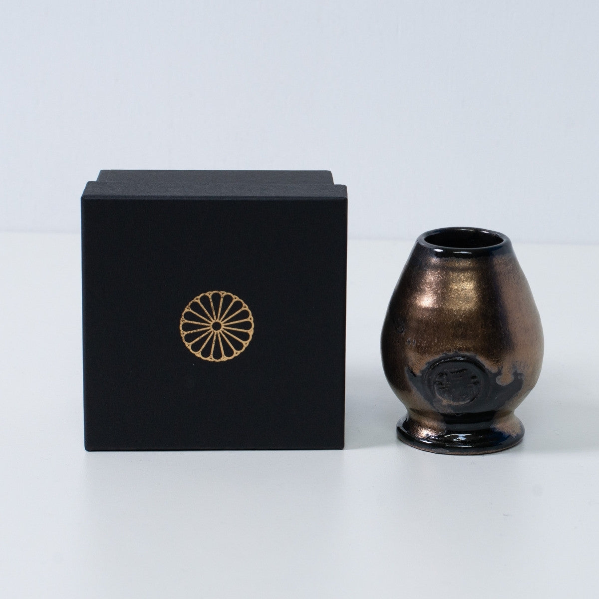 Gold and black Japanese matcha whisk stand "Zen" pottery next to a black gift box with a gold chrysanthemum crest, from Japan.