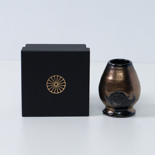 Gold and black Japanese matcha whisk stand "Zen" pottery next to a black gift box with a gold chrysanthemum crest, from Japan.