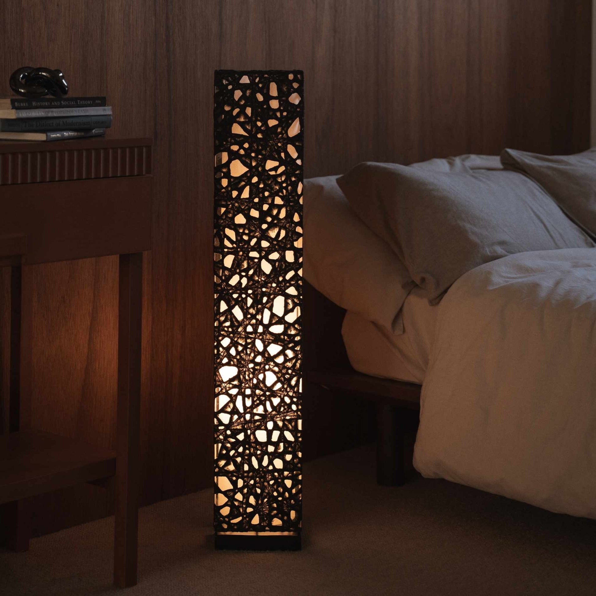 Lit black Japanese paper floor lamp Me Look 01 standing next to a bed in a dim japanese bedroom context.