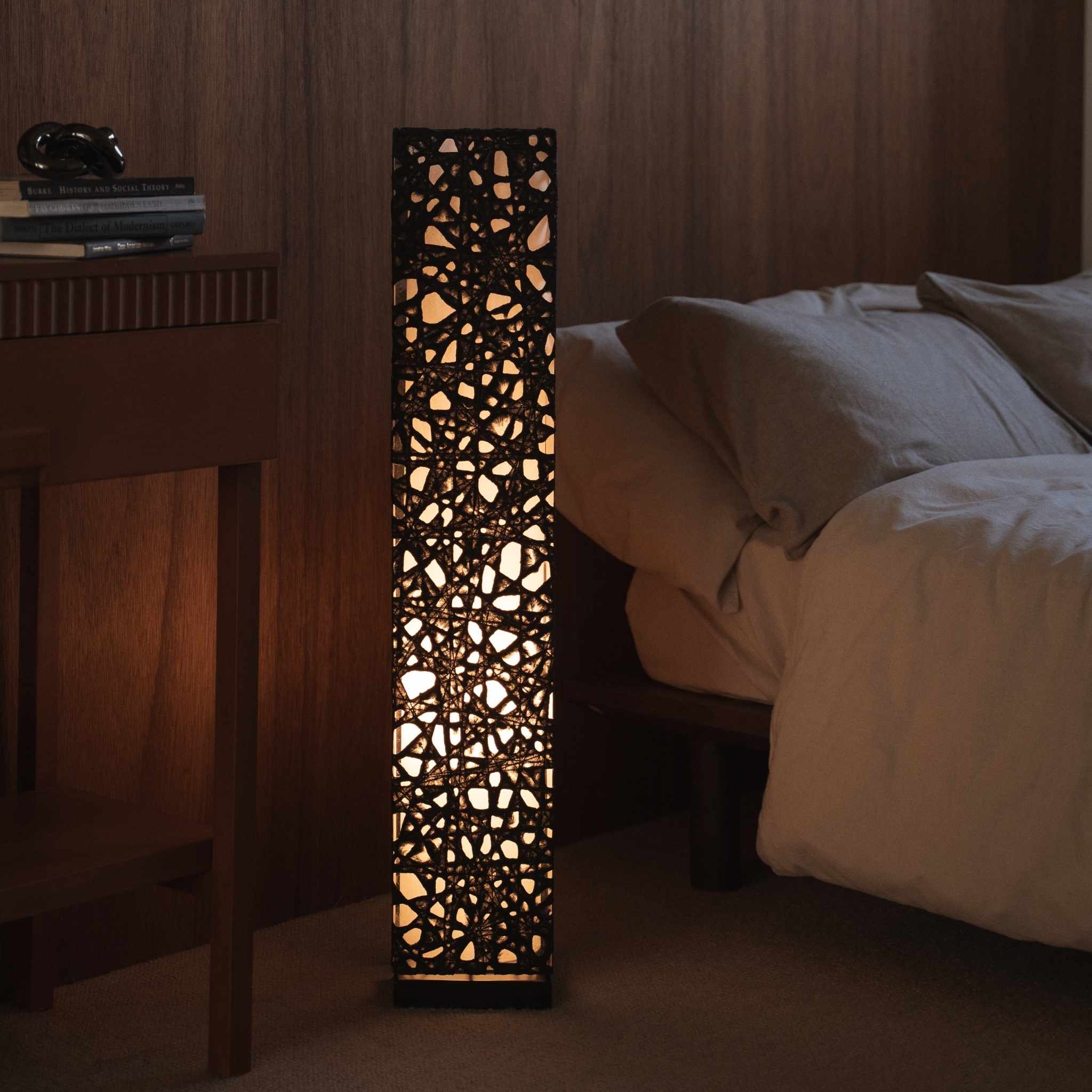 Lit black Japanese paper floor lamp Me Look 01 standing next to a bed in a dim japanese bedroom context.