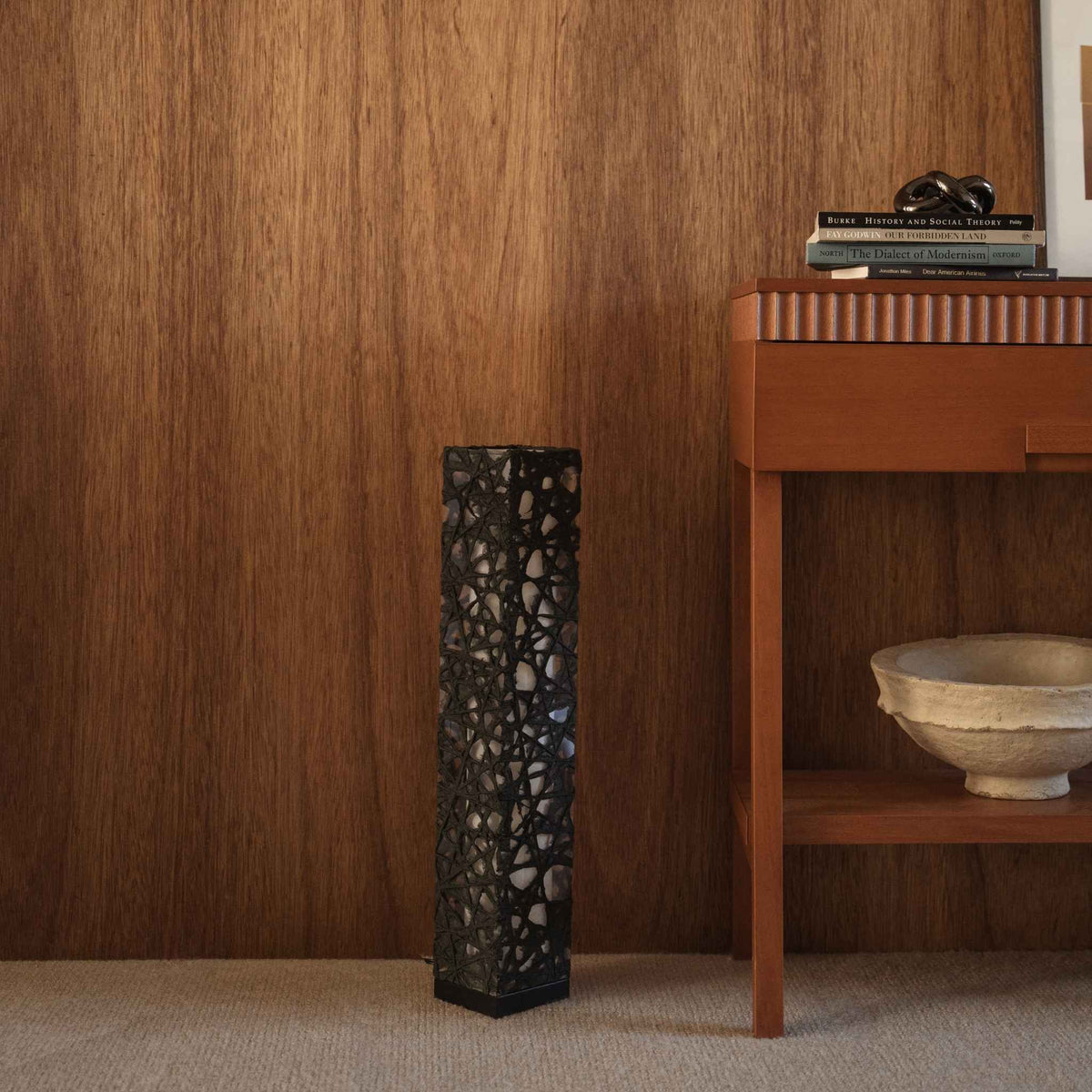 Black Me Look 02 paper floor lamp standing unlit on a carpet against a dark wood wall near a console table.