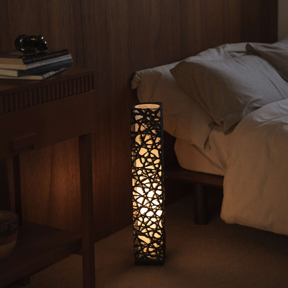 Lit black Japanese paper floor lamp Me Look 02 glowing warmly beside a bed in a japanese bedroom context.