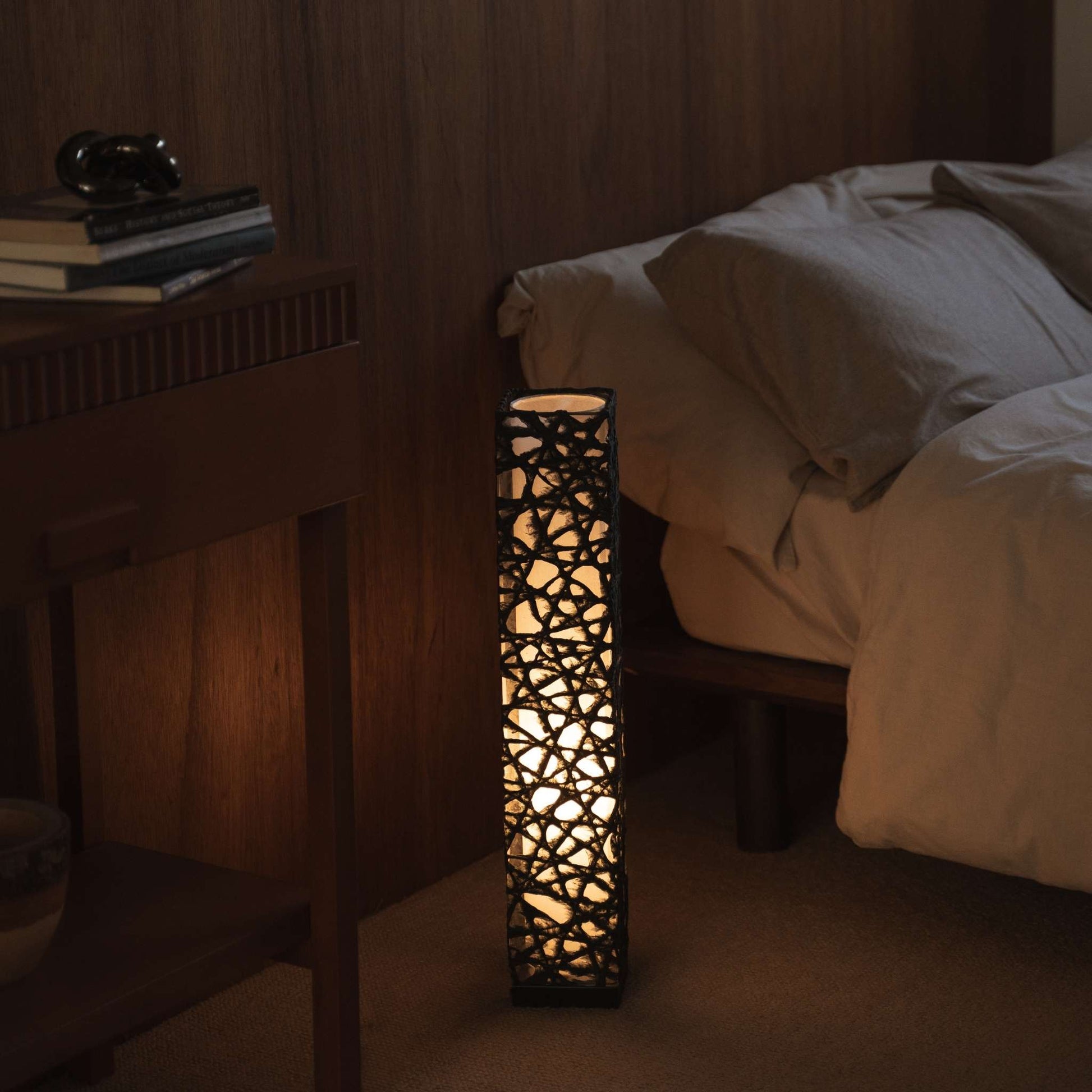 Lit black Japanese paper floor lamp Me Look 02 glowing warmly beside a bed in a japanese bedroom context.