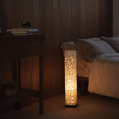 Lit white Japanese paper floor lamp Me Look 02 glowing warmly beside a bed in a japanese bedroom context.