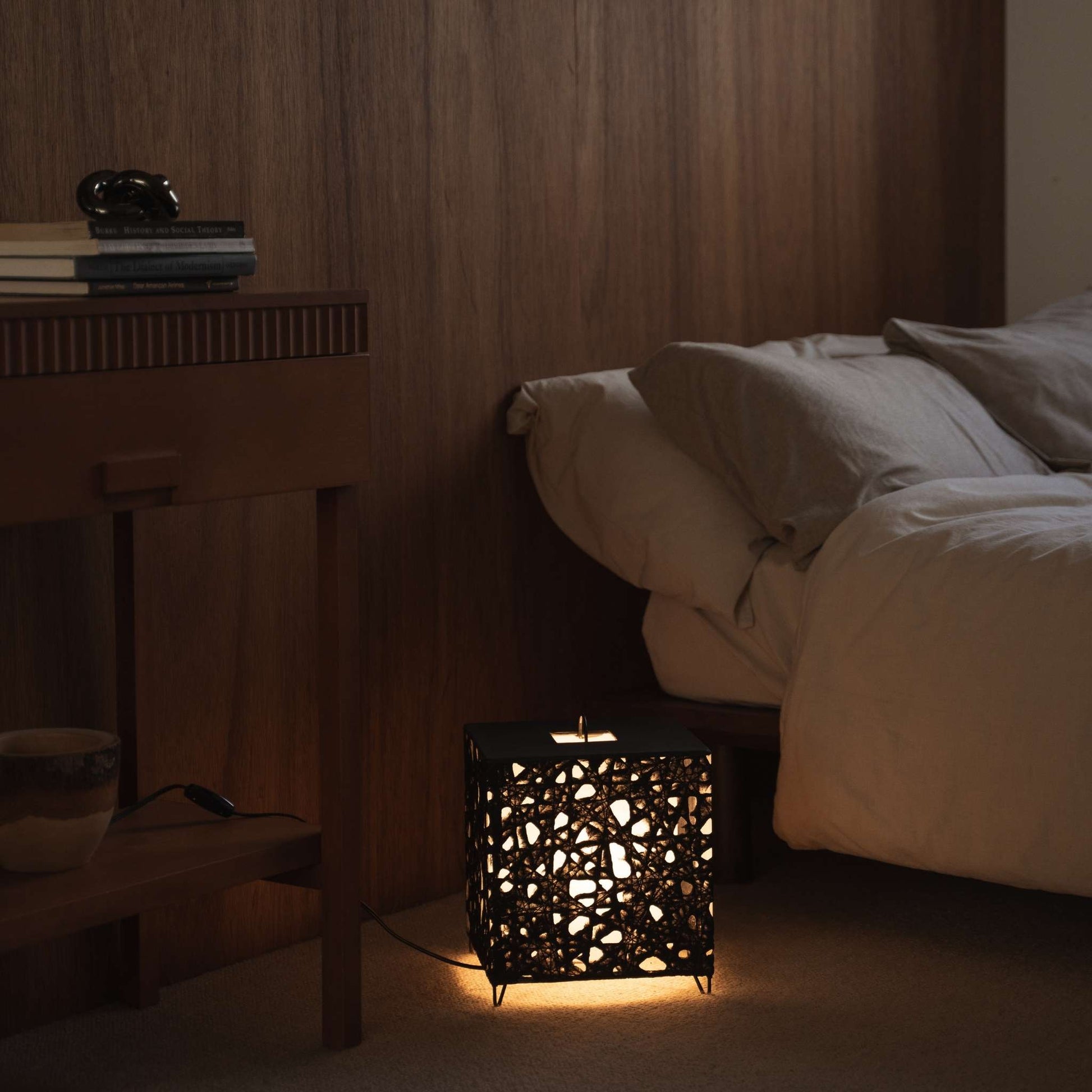 Lit cube shaped Japanese paper lamp Me-Look 03 black glowing warmly on a wooden bedside table in a japanese bedroom.