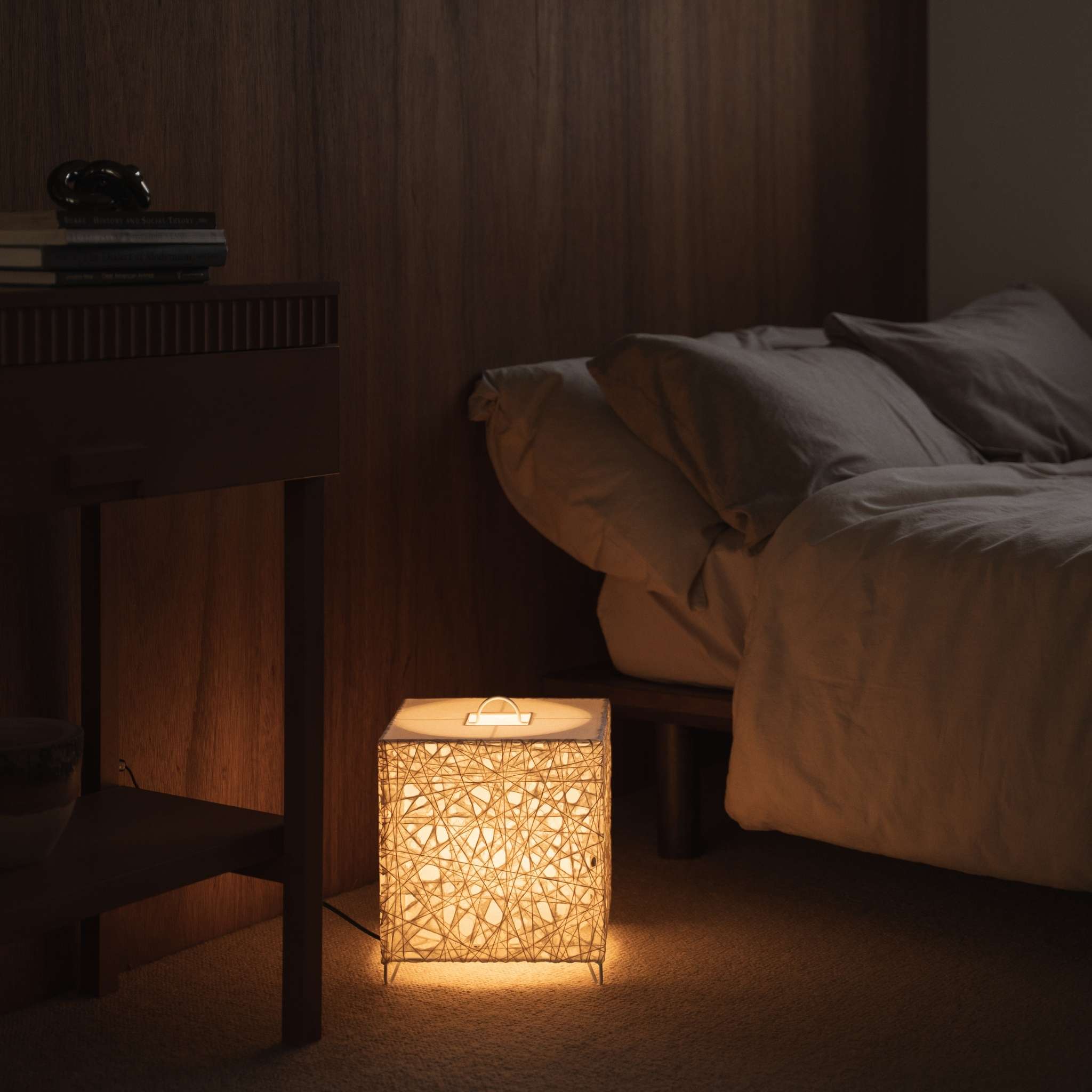 Lit cube shaped Japanese paper lamp Me-Look 03 white glowing warmly on a wooden bedside table in a Japanese bedroom.