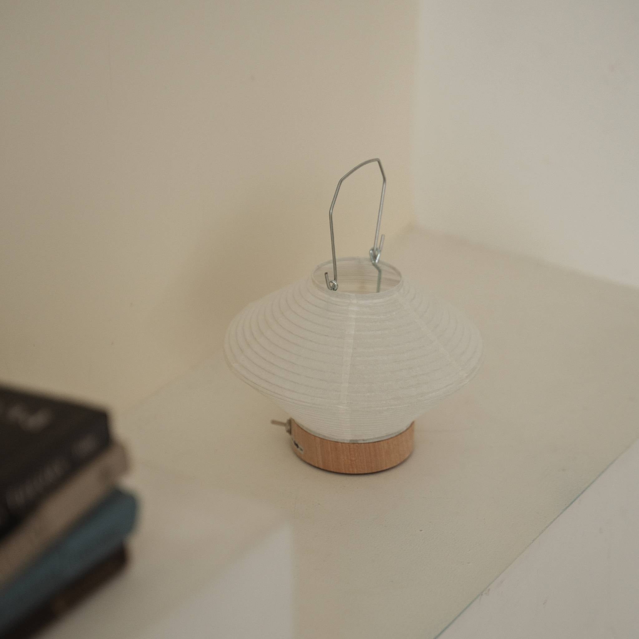 A portable japanese paper lamp RIN 05 with a metal handle sitting on a white surface showing its delicate mino washi texture