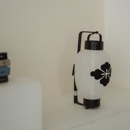 Side profile of a standing paper lantern showing the black bamboo bow structure against a white wall.