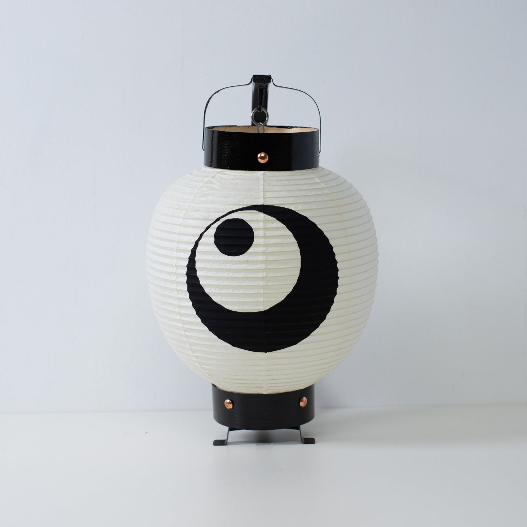 A small Japanese paper lantern with a hand-painted black moon and star crest standing on a white surface.