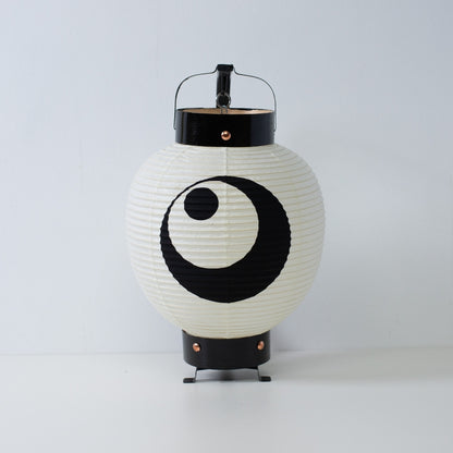 A small Japanese paper lantern with a hand-painted black moon and star crest standing on a white surface.