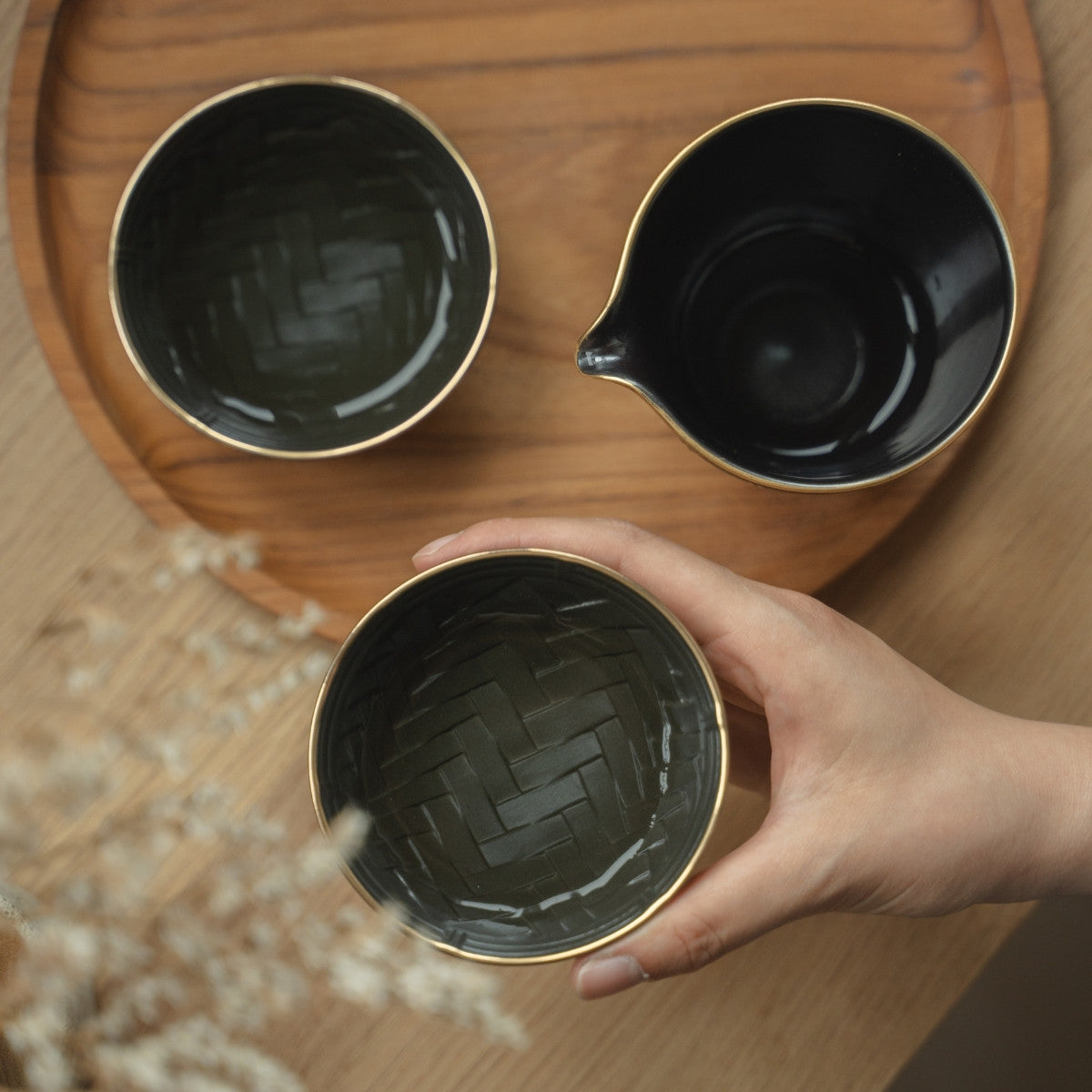 Hand holding a black and gold sake cup showing the basket weave texture over a tray, preparing for a mindful evening.