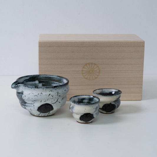 Weißes und schwarzes japanisches Sake-Set "Creative Cloud" aus Keramik mit einer Sake-Karaffe und zwei Bechern, neben einer Holzkiste mit Chrysanthemen-Emblem.