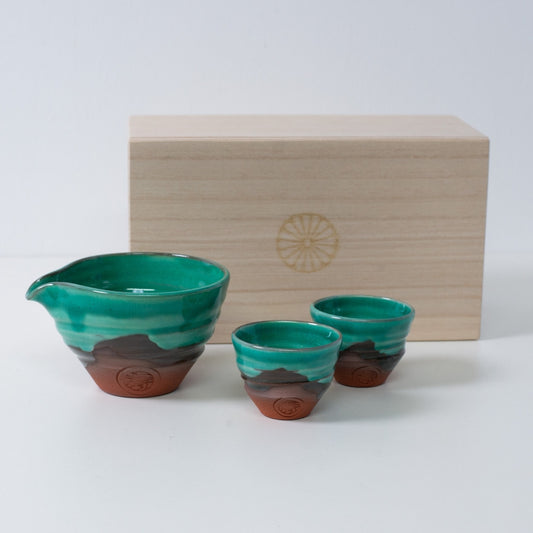 Sake-Set "Emerald" aus Keramik in Smaragdgrün und Orange, japanisch, mit Sake-Karaffe und zwei Bechern, neben einer Holzbox mit Chrysanthemen-Wappen.
