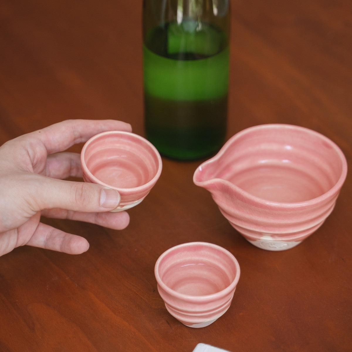 Eine Person hält einen pinken japanischen Kyo-yaki Sakebecher (Ochoko) über einen Holztisch, daneben eine Sakeflasche und eine Katakuchi-Karaffe.