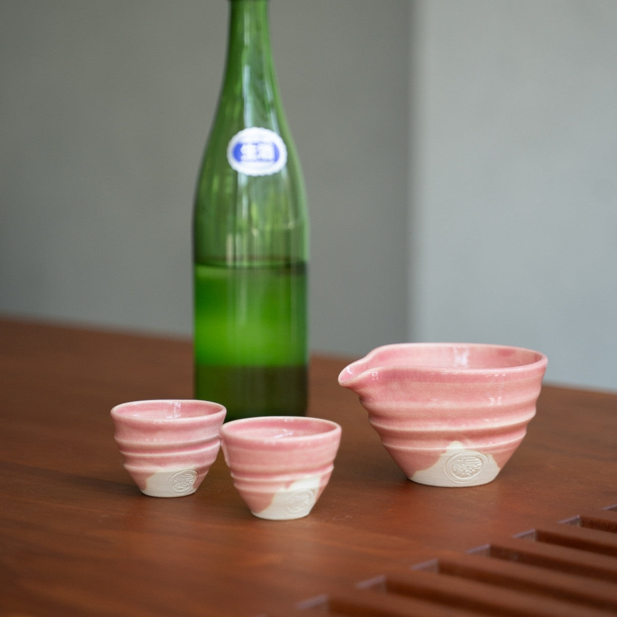 Pinkes japanisches Kyo-yaki Keramik-Sake-Set (Katakuchi-Karaffe, zwei Becher) auf einem Holztisch im Japandi-Stil, bereit für ein besonderes Abendessen.