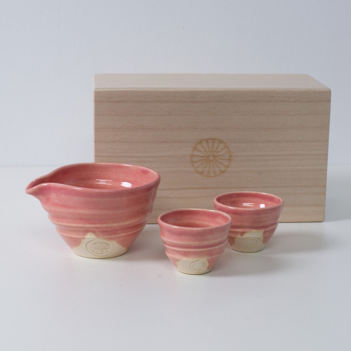 Pinkes japanisches Sake-Set "Omuro Cherry" aus Keramik mit einer Sake-Karaffe und zwei Bechern, neben einer Holzkiste mit Chrysanthemen-Wappen.