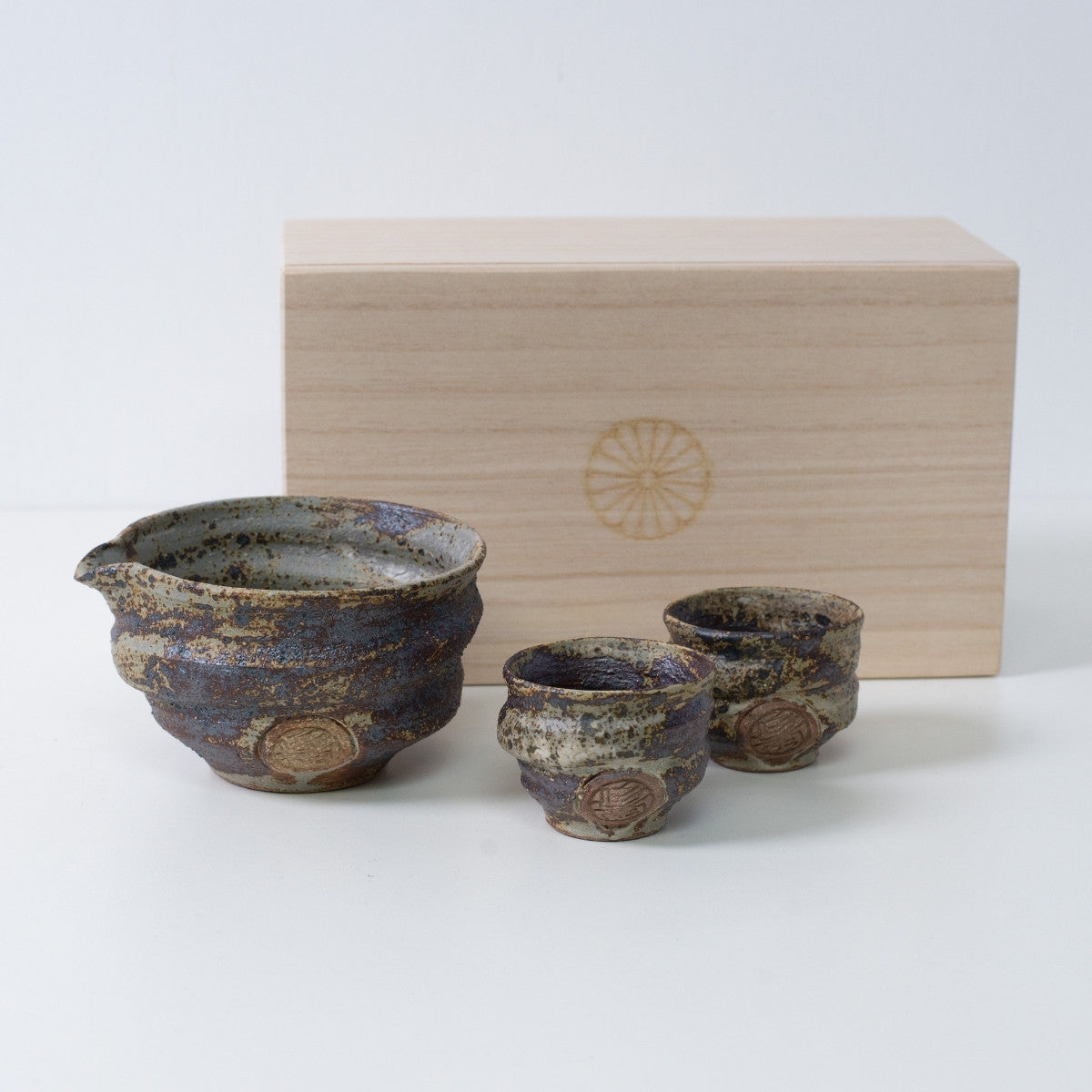 Rustikales braunes japanisches Sake-Set "Sakigake" aus Keramik mit einer Sake-Karaffe und zwei Bechern, neben einer Holzkiste mit Chrysanthemen-Emblem.