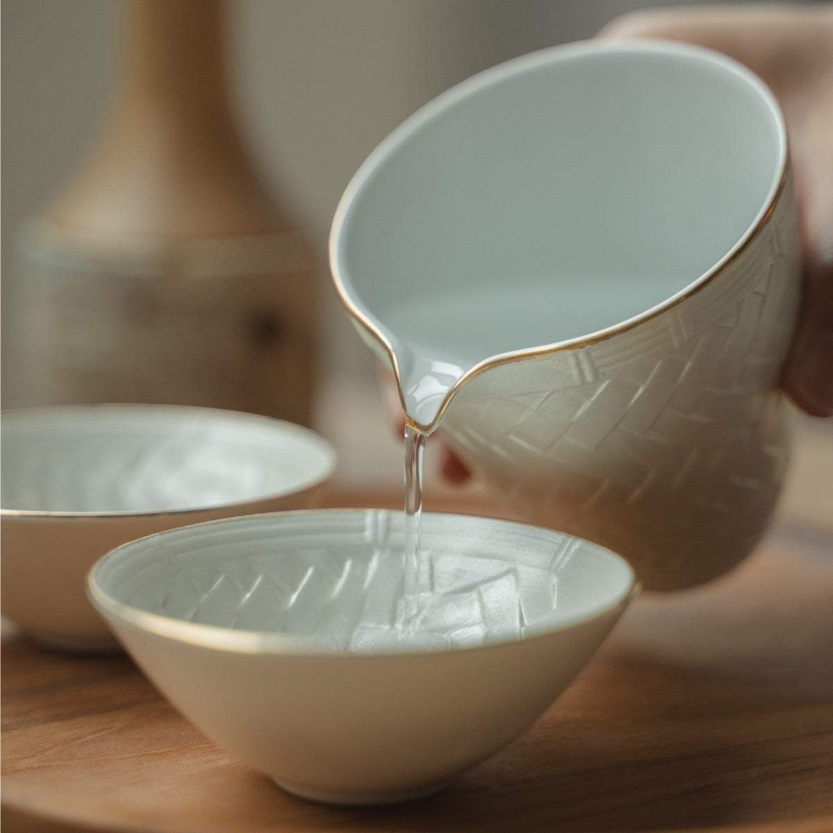 Hand pouring from a white Japanese sake pitcher into a delicate guinomi cup on a wooden tray, elevating a special dinner.