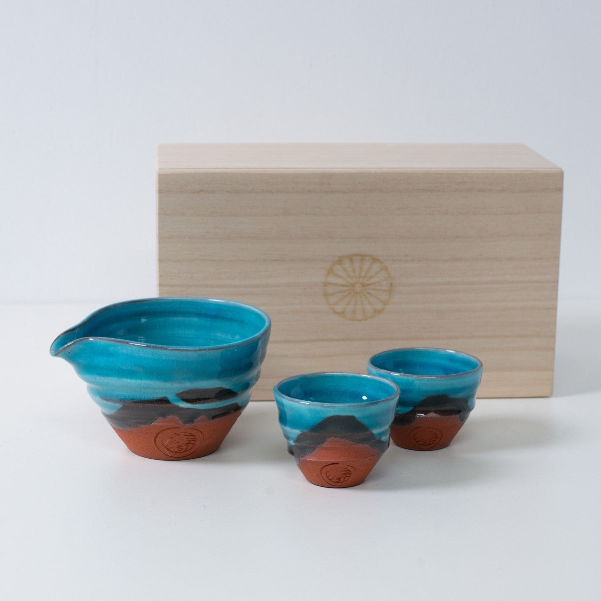 Türkisfarbenes japanisches Sake-Set "Soku Blue" aus Keramik mit Sake-Karaffe und zwei Bechern, neben einer Holzkiste mit Chrysanthemen-Emblem.