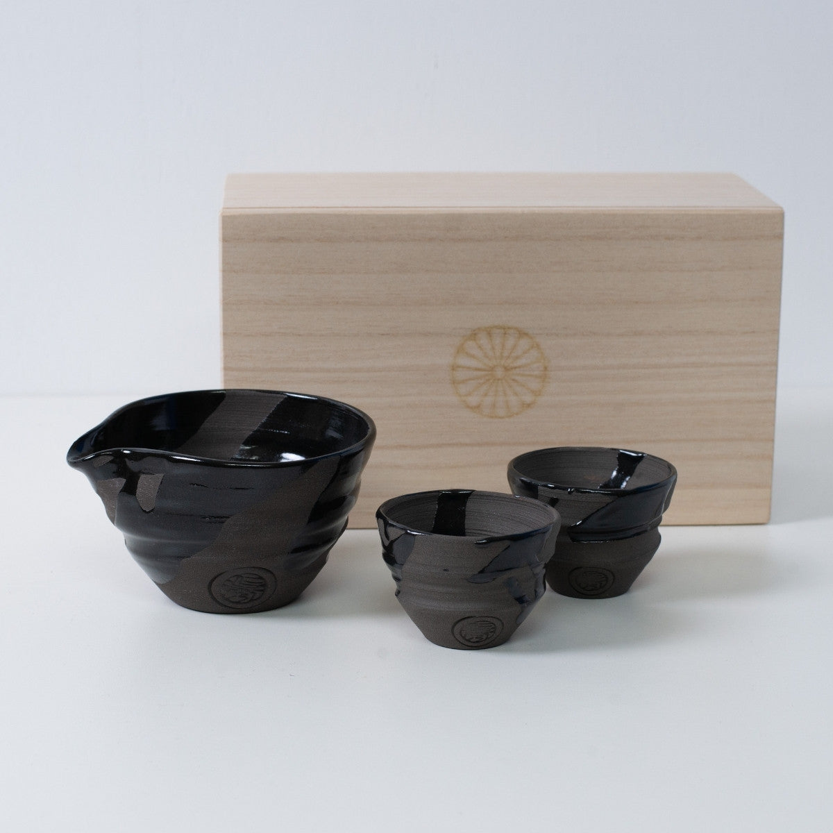 Japanisches Sake-Set „Sumi Black“ aus Keramik mit Sake-Karaffe und zwei Bechern, neben einer Holzkiste mit Chrysanthemen-Emblem.