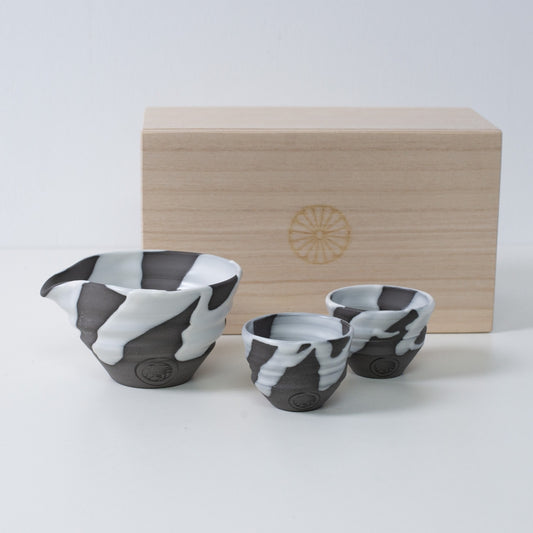 Weiß-graues japanisches Sake-Set „White Flow“ aus Keramik mit Sake-Karaffe und zwei Bechern, neben einer Holzkiste mit Chrysanthemen-Emblem.