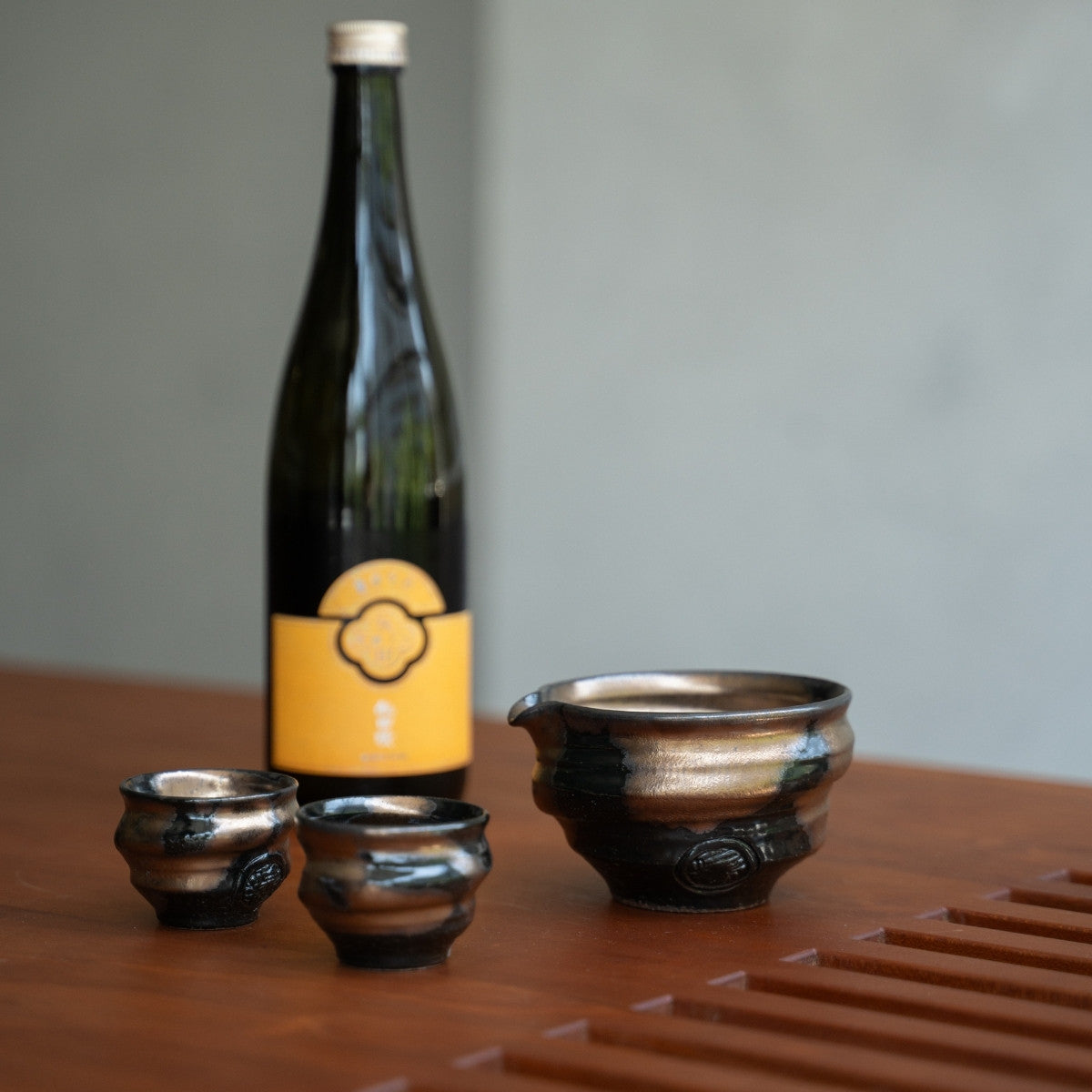 Japanese Sake Set (Kyo-yaki, gold/black) and a bottle arranged on a minimalist wooden table, creating a serene, inviting scene.