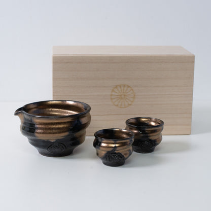 Gold-schwarzes japanisches Sake-Set „Zen“ aus Keramik mit einer Karaffe und zwei Bechern neben einer Holzkiste mit Chrysanthemen-Wappen.
