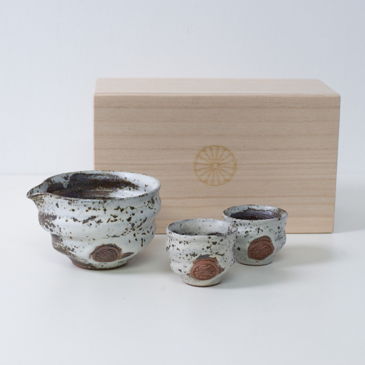 Braunes und weißes japanisches Sake-Set "Zui" aus Keramik mit Sake-Karaffe und zwei Bechern, neben einer Holzkiste mit Chrysanthemen-Wappen.