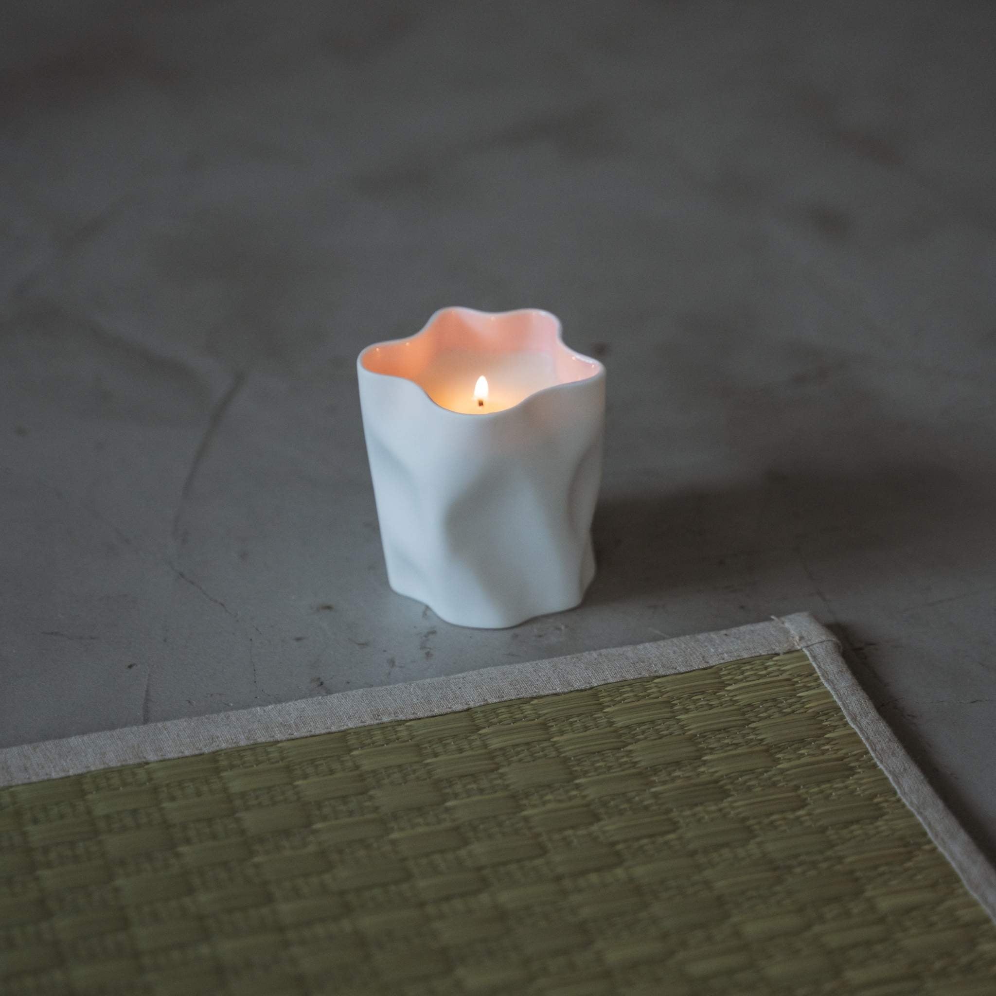 Bougie parfumée à la fleur de cerisier dans un récipient en porcelaine blanche, illuminée d'une douce lumière rose dans un décor zen paisible.