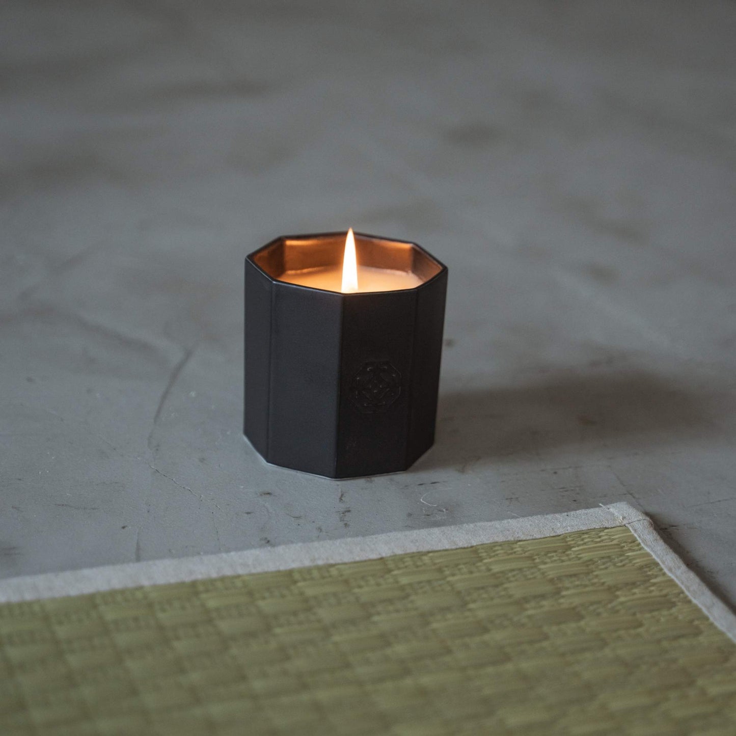 Black arita ware scented candle burning with a warm flame on a grey floor next to a traditional tatami mat.