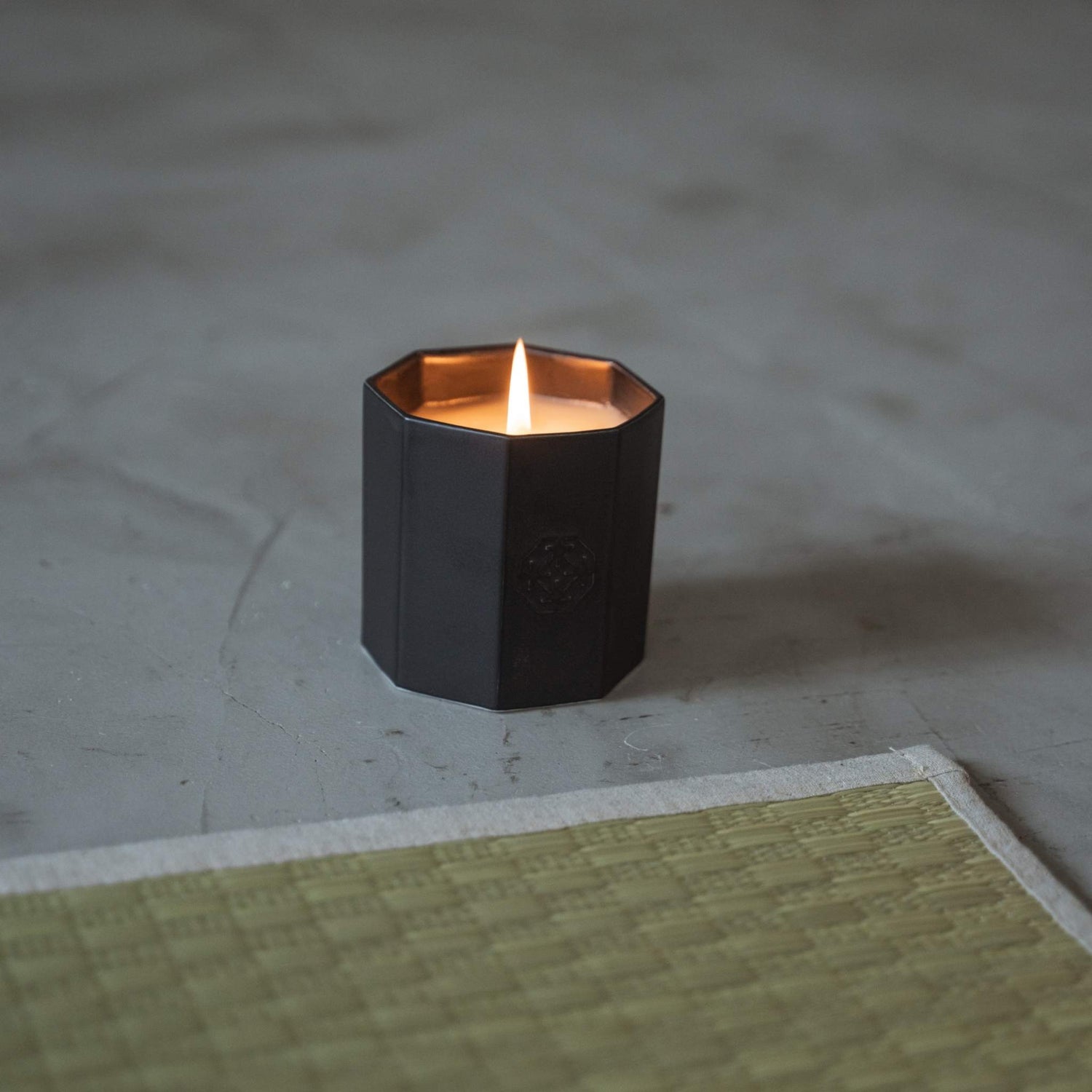 Black arita ware scented candle burning with a warm flame on a grey floor next to a traditional tatami mat.