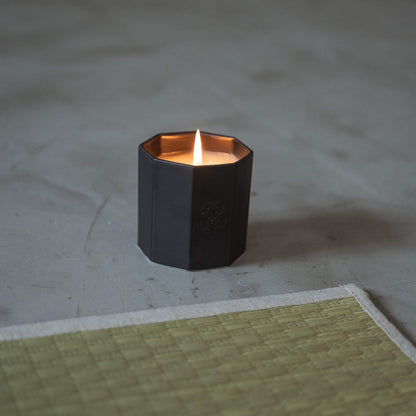 Black arita ware scented candle burning with a warm flame on a grey floor next to a traditional tatami mat.