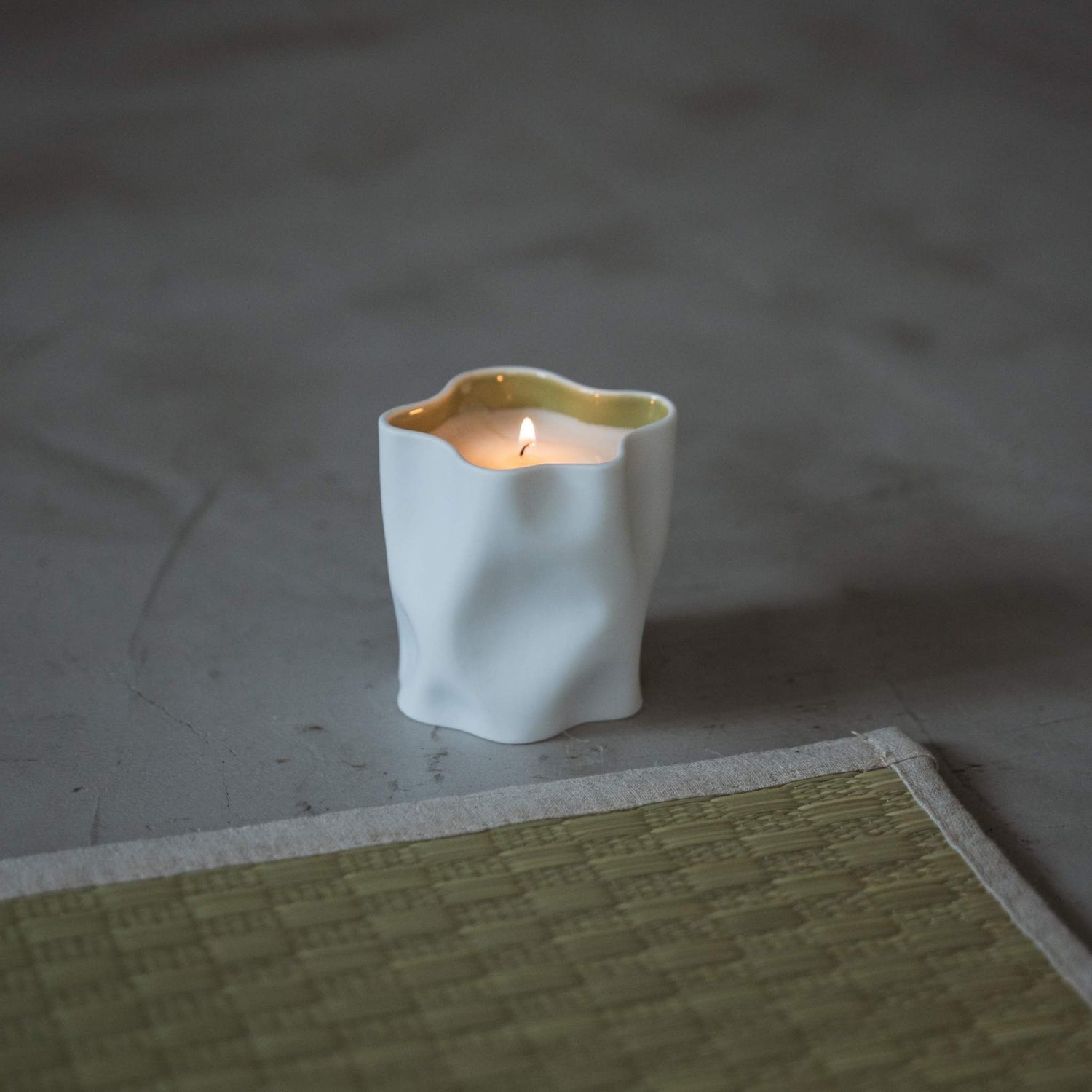 Yuzu japanese scented candle in a white porcelain vessel burning with a soft flame next to a tatami mat.