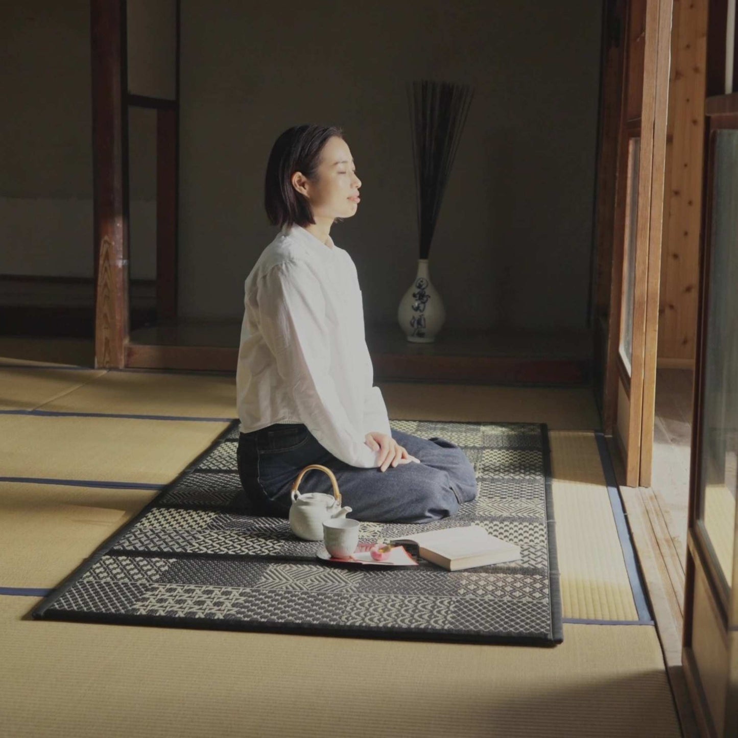 Woman meditating on a black patterned tatami mat with a tea set creating a serene zen decor space.