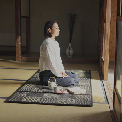 Woman meditating on a black patterned tatami mat with a tea set creating a serene zen decor space.