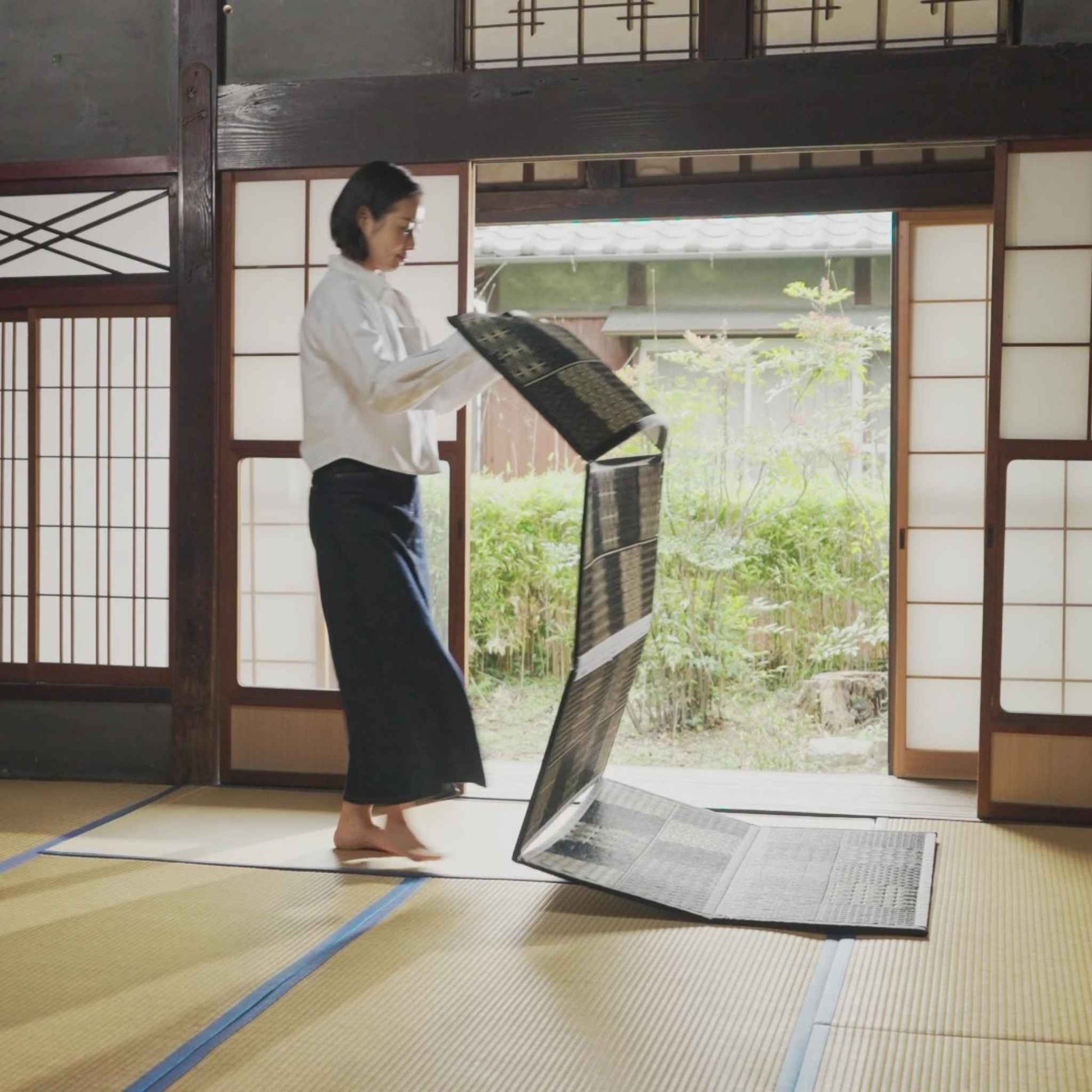 Woman unfolding a black five panel tatami mat on a traditional floor preparing a japanese decor space.