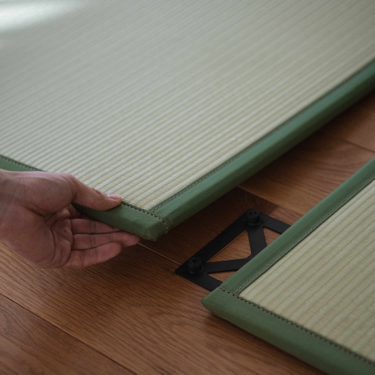 Hands connecting two green tatami mat panels on a floor demonstrating easy setup for a custom Japanese space.