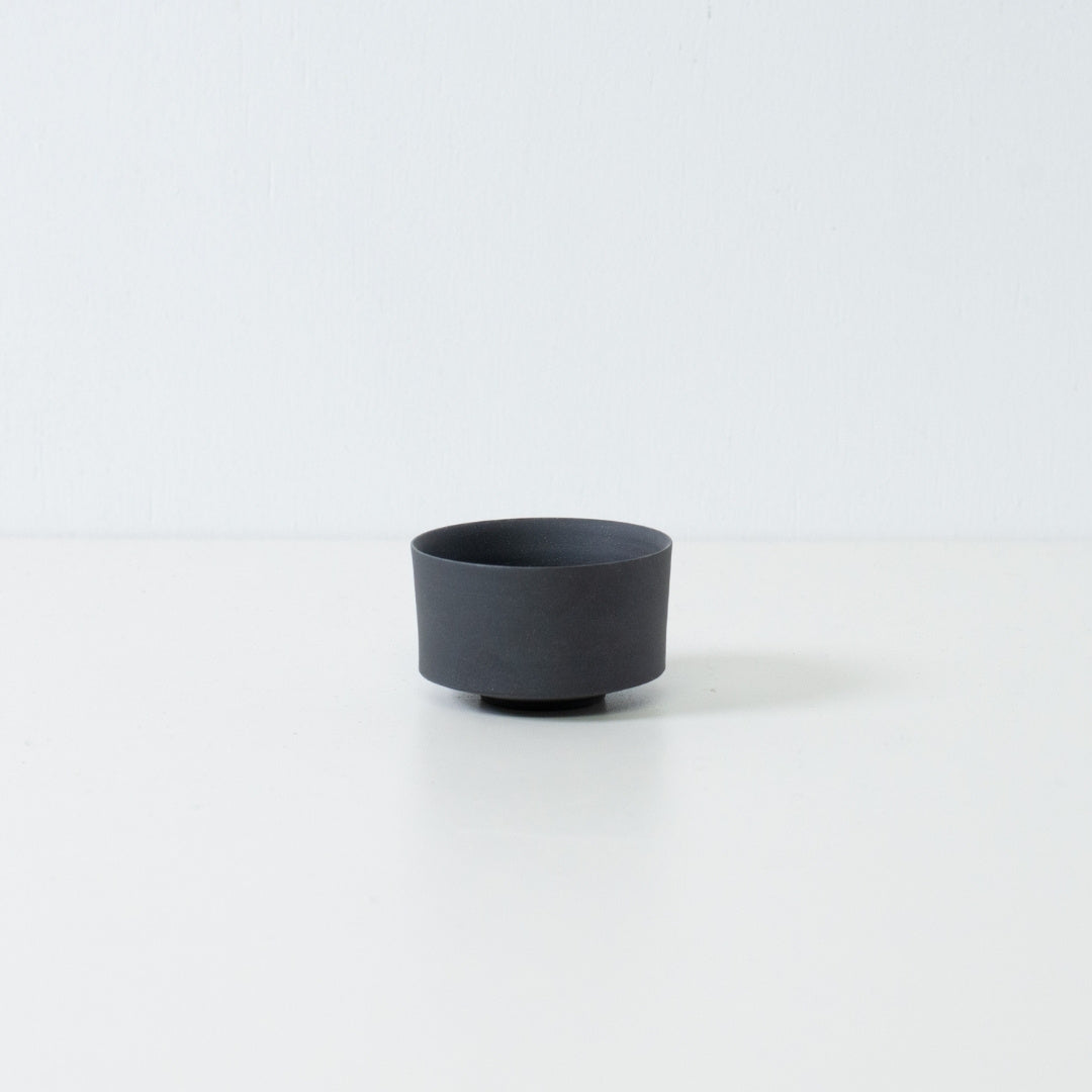 Single matte black japanese tea cup with a straight cylinder shape and stone like texture sitting on a white surface