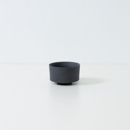 Single matte black japanese tea cup with a straight cylinder shape and stone like texture sitting on a white surface