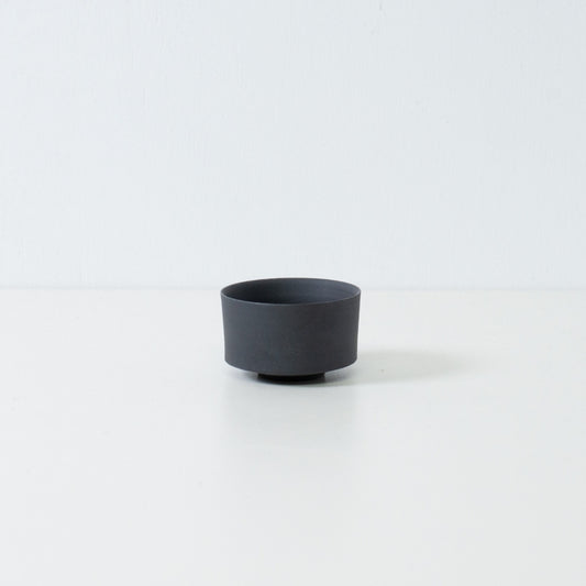 Single matte black japanese tea cup with a straight cylinder shape and stone like texture sitting on a white surface