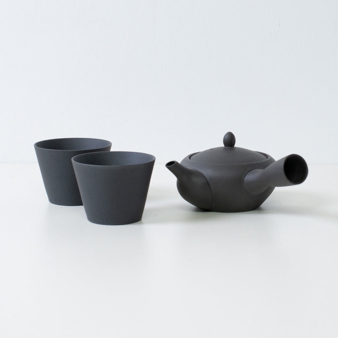 Matte black japanese tea set featuring a monks bowl kyusu teapot and stackable tea cups on a white table.