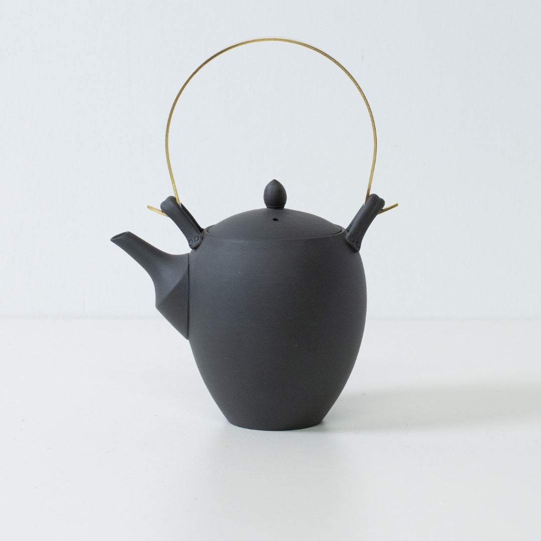Side view of a black japanese teapot with an acorn shape and brass handle showcasing traditional banko ware craftsmanship