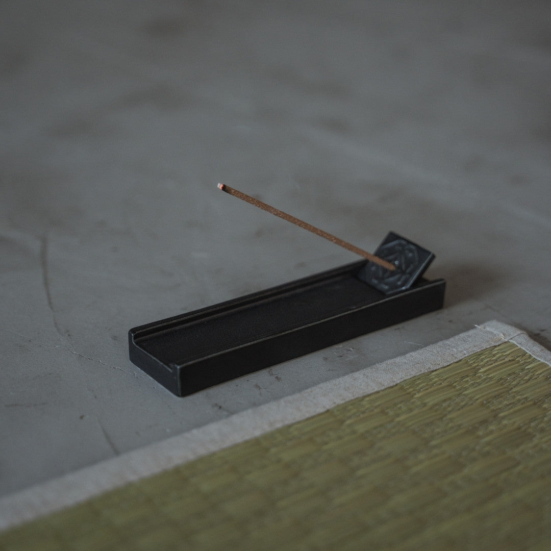 A Japanese Hinoki incense stick burns in a black porcelain holder on a zen tatami yoga mat for wellness.