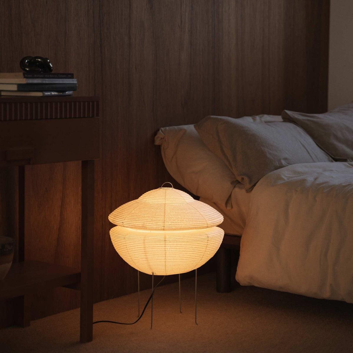 Illuminated Japanese paper floor lamp Gemini 01 glowing with warm soft light situated on the floor beside a bed in a Japanese bedroom