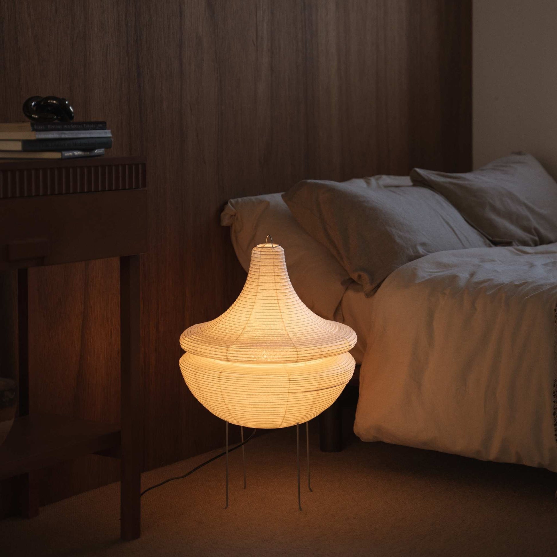 Illuminated Japanese paper floor lamp Gemini 02 glowing with warm soft light situated on the floor beside a bed in a Japanese bedroom
